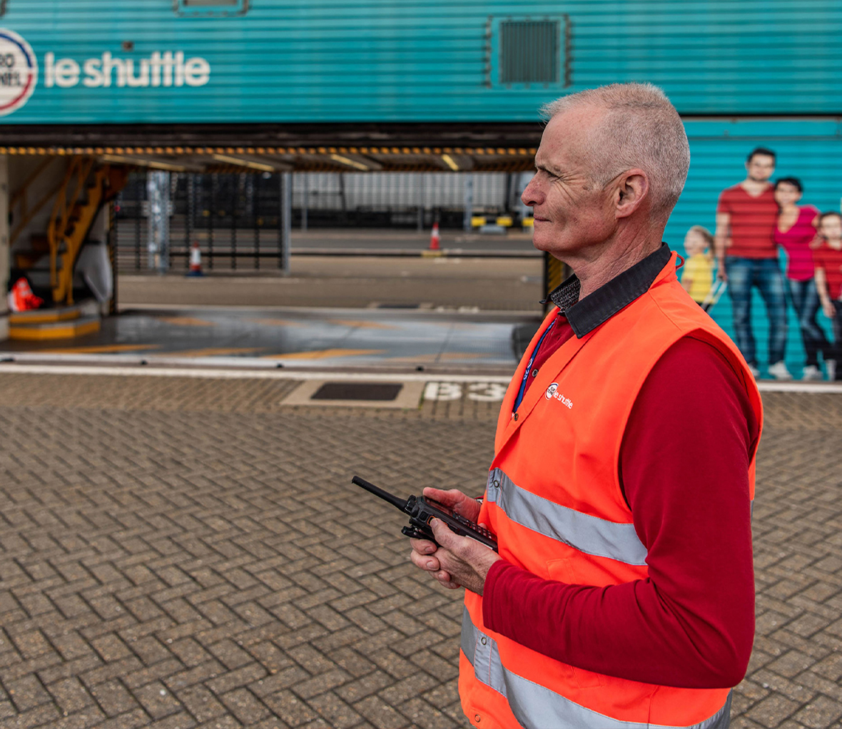 Grey haired man holding a walkie talkie wearing a red long sleeve top and bright orange vest standing in front of a blue Eurotunnel shuttle
